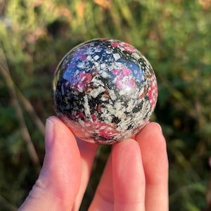 May include: A polished spherical stone featuring a mottled pattern of deep black, bright magenta, and off-white crystalline inclusions. The surface is smooth and reflective, showcasing a complex mineral composition with distinct colour variations.