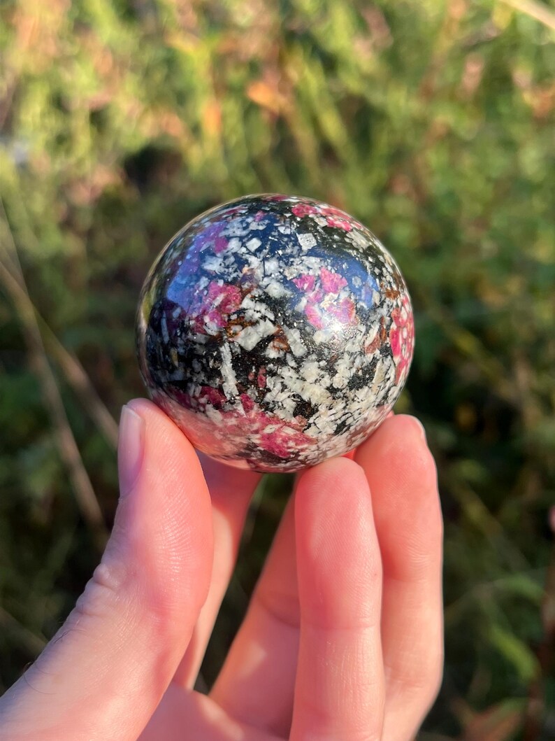 May include: A polished spherical stone featuring a mottled pattern of deep black, bright magenta, and off-white crystalline inclusions. The surface is smooth and reflective, showcasing a complex mineral composition with distinct colour variations.