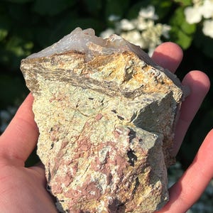 May include: A natural rock formation with a crystalline top. The rock displays layers of tan, green, and reddish-pink hues. The stone is held in a hand, with a blurred green and white floral background.