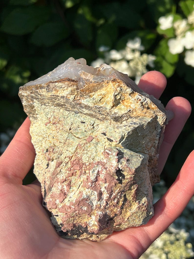 May include: A natural rock formation with a crystalline top. The rock displays layers of tan, green, and reddish-pink hues. The stone is held in a hand, with a blurred green and white floral background.