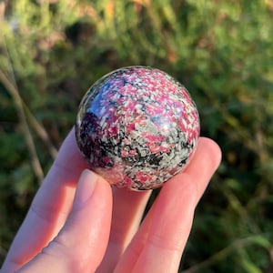 May include: A polished spherical stone featuring a mottled pattern of vibrant magenta, white, and black mineral inclusions. The surface of the sphere is smooth and reflective, showcasing a complex crystalline texture throughout the entire round object.