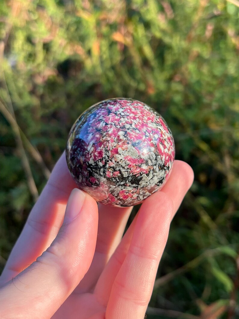 May include: A polished spherical stone featuring a mottled pattern of vibrant magenta, white, and black mineral inclusions. The surface of the sphere is smooth and reflective, showcasing a complex crystalline texture throughout the entire round object.