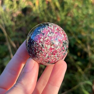 May include: A polished spherical stone featuring a vibrant mix of deep magenta, white, and black mineral inclusions. The surface is smooth and reflective, showcasing a complex, speckled pattern of contrasting colors throughout the entire round object.
