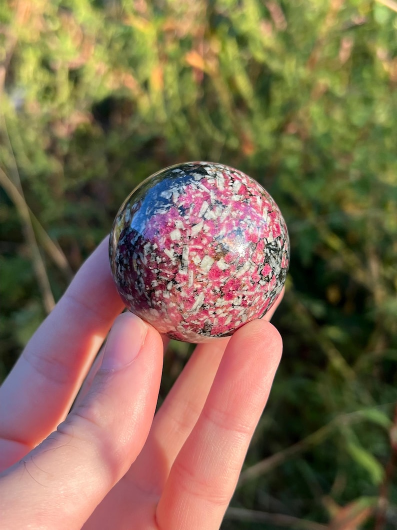 May include: A polished spherical stone featuring a vibrant mix of deep magenta, white, and black mineral inclusions. The surface is smooth and reflective, showcasing a complex, speckled pattern of contrasting colours throughout the entire round object.