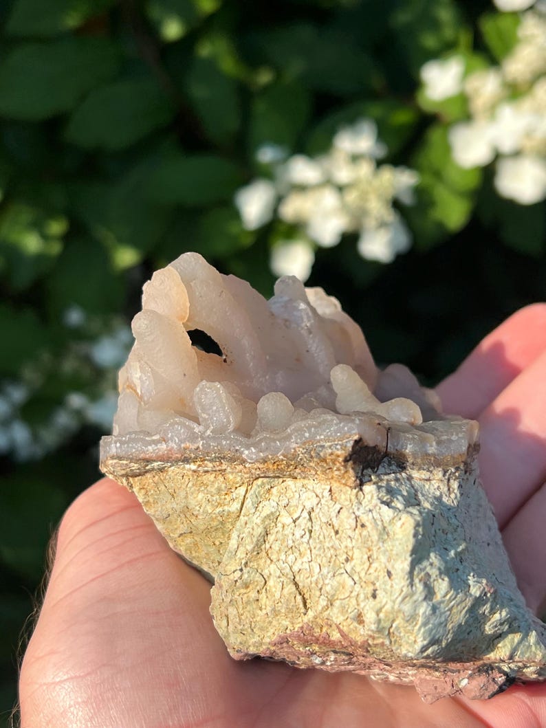 May include: A light-coloured, crystalline mineral specimen with a rough, textured base. The top portion features a cluster of translucent, rounded formations. The base is a mix of tan and green hues, with visible cracks and imperfections.