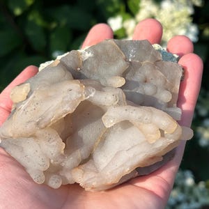 May include: A light grey and beige mineral specimen with a bubbly, layered texture. The stone is held in a hand, with a blurred green and white background. The mineral has a translucent quality, with some areas appearing more opaque.