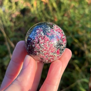 May include: A polished spherical stone featuring a mottled pattern of vibrant pink, white and black mineral inclusions. The surface is smooth and reflective, showcasing a dense, crystalline texture with varied colour distribution throughout the sphere.
