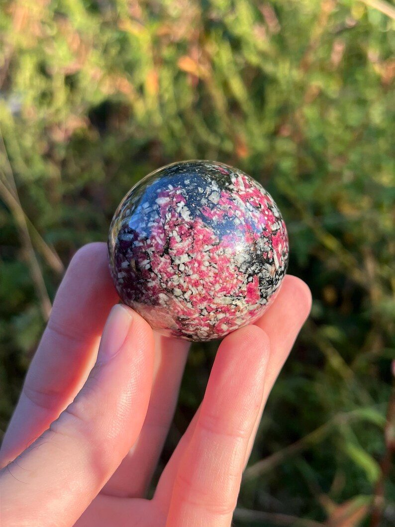 May include: A polished spherical stone featuring a mottled pattern of vibrant pink, white and black mineral inclusions. The surface is smooth and reflective, showcasing a dense, crystalline texture with varied colour distribution throughout the sphere.