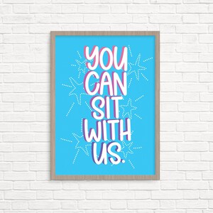Op de afbeelding: Een ingelijst print met een blauwe achtergrond en witte tekst die "YOU CAN SIT WITH US." luidt. De tekst is in een vet, speels lettertype met een roze omtrek. De achtergrond is versierd met witte sterren.