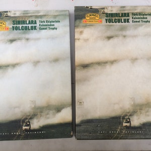 Op de afbeelding: Twee boekomslagen met een groen-geel kleurenschema. De omslagen tonen een foto van een auto die over een stoffige weg rijdt met de tekst "Camel Trophy" en "Sinirlara Yolculuk" in wit. De tekst onder de foto luidt "Turk Ekiplerinin Kaleminden Camel Trophy".