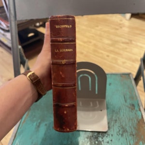 May include: A vintage, brown leather-bound book with gold lettering on the spine. The title is "Bromfield" and "La Mousson". The book is standing upright in a silver bookend.