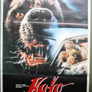 May include: Movie poster for "Cujo" featuring a menacing, snarling dog with exposed teeth and blood. A car with terrified occupants is visible. The title "Cujo" is in red, with the names of the actors below.