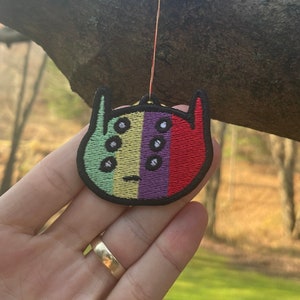 May include: A colourful embroidered patch with a cartoon face and rainbow stripes. The patch has a green, yellow, purple, and red stripe. The face has black eyes and a black mouth.
