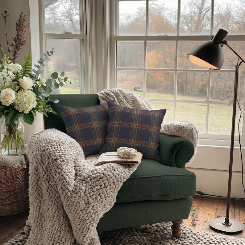 K&ouml;nnte beinhalten: Ein gem&uuml;tlicher Sessel in Dunkelgr&uuml;n mit zwei karierten Kissen. Eine dicke Strickdecke und eine Vase mit wei&szlig;en Blumen sind zu sehen. Eine schwarze Stehlampe und ein Fenster mit Aussicht vervollst&auml;ndigen die Szene.