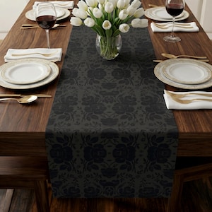 May include: A dark gray table runner featuring a repeating black floral pattern laid across a wooden dining table. The table is set with white plates, gold-toned silverware, white napkins, wine glasses, and a glass vase filled with white tulips.