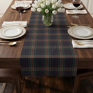 May include: A rectangular table runner featuring a dark navy blue, forest green, and thin red plaid pattern. The fabric runner is centred on a wooden table, providing a base for a clear glass vase filled with white tulips and place settings.
