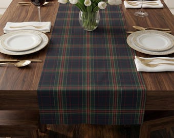 May include: A rectangular table runner featuring a dark navy blue, forest green, and thin red plaid pattern. The fabric runner is centered on a wooden table, providing a base for a clear glass vase filled with white tulips and place settings.