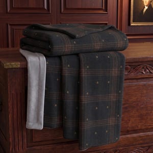 May include: A folded charcoal gray blanket featuring a subtle brown plaid pattern and small embroidered gold crests. The blanket has a soft, light gray underside visible along the edge, showcasing a dual-textured design with a smooth finish.