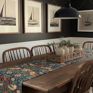 May include: A dining room with a dark wood table, a patterned table runner, and wooden chairs. Framed sailboat art hangs on the dark wall. A woven basket with vases and greenery sits on the table.