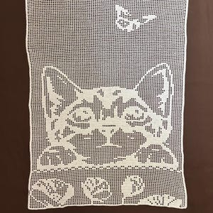May include: White crocheted rectangular doily featuring a cat face design. The cat's face is centered, with detailed features and a butterfly above. The doily is made of a fine, open weave, and is likely handmade.