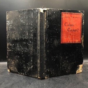 May include: A black hardcover book with a red label that reads "Cuban Cookery".