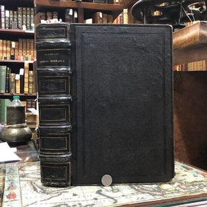 May include: A large, black leather-bound book with gold accents and the title "Biblia Hebraica" on the spine. The book is sitting on a table with a map-like surface.