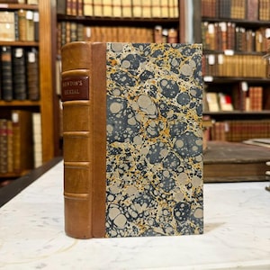 May include: Antique book titled "Newton's Herbal" with a brown leather spine and marbled cover in shades of gray, black, and gold. The book is displayed on a white surface in a library setting, surrounded by other books.