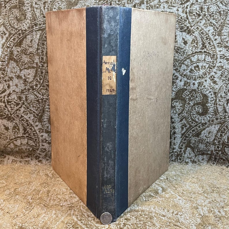 Puede incluir: Un libro antiguo de tapa dura con una encuadernaci&oacute;n azul oscuro y una cubierta de color beige. La encuadernaci&oacute;n tiene el texto "Aerial Age" y "1925" impreso.