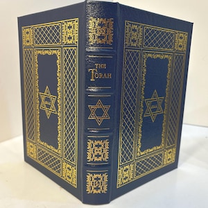 May include: A dark blue hardcover book titled The Torah with a textured finish. The cover and spine feature intricate gold foil stamping, including geometric lattice patterns, ornate floral borders, and a Star of David with Hebrew text inside.