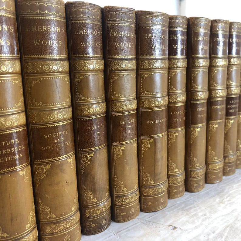 May include: A collection of antique books with brown leather covers and gold lettering. The spines display titles such as "Emerson's Works" and "Essays." Decorative gold filigree accents the spines and covers, creating a classic aesthetic.