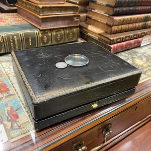 May include: A black leather box with gold trim and a magnifying glass on top. The box is sitting on a wooden table with a drawer.
