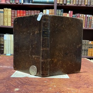 May include: A brown leather-bound book with the title "DE" and "COR" visible on the spine. The book is sitting on a wooden surface.