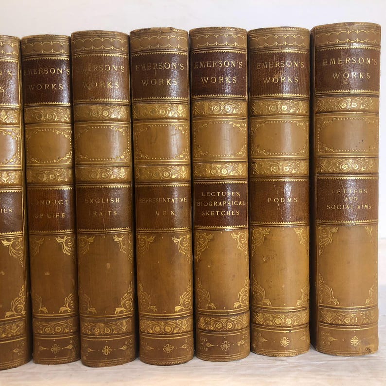 May include: A collection of antique books with brown leather covers and gold embossed lettering and designs. The spines read "Emerson's Works" and feature titles like "Conduct of Life" and "English Traits."