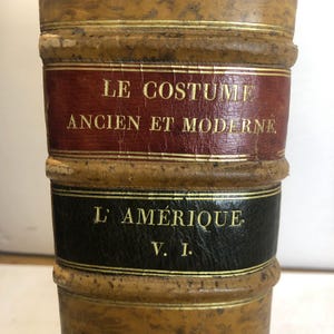 May include: Close-up of the spine of two antique books. The top book has a burgundy label with gold lettering that reads "LE COSTUME ANCIEN ET MODERNE." The bottom book has a black label with gold lettering that reads "L'AMERIQUE V. I."