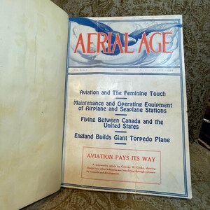 Puede incluir: Una portada de revista vintage para "Aerial Age" de enero de 1922. La portada presenta una ilustraci&oacute;n azul y blanca de un avi&oacute;n en vuelo. El texto "Aviation and The Feminine Touch" est&aacute; impreso en negro sobre un fondo blanco.