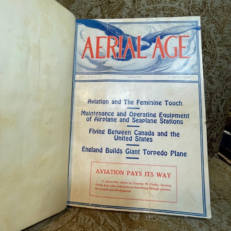Puede incluir: Una portada de revista vintage para "Aerial Age" de enero de 1922. La portada presenta una ilustraci&oacute;n azul y blanca de un avi&oacute;n en vuelo. El texto "Aviation and The Feminine Touch" est&aacute; impreso en negro sobre un fondo blanco.