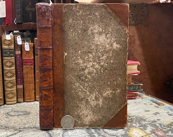 May include: A vintage, brown leather-bound book with a worn, gray cover. The book has a decorative spine with gold lettering and a metal clasp. The book is sitting on a map.