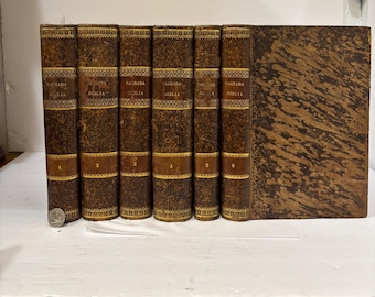 May include: A collection of antique books with brown leather covers. The spines display gold lettering and decorative patterns. The books are numbered and titled "Sagrada Biblia". The covers have a marbled design.