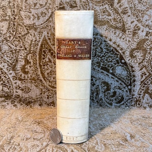 May include: A vintage book titled "Cary's Great Roads of England & Wales" with a brown leather spine and gold lettering. The book is bound in white paper and has a date of 1817 printed on the bottom.