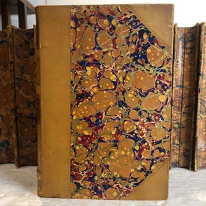 May include: A collection of antique books with marbled covers. The covers feature a tan leather spine and corners, with a decorative pattern of swirling gold, brown, blue, and red. The books are stacked together, showcasing their aged appearance.