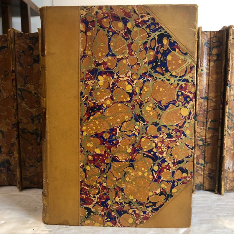 May include: A collection of antique books with marbled covers. The covers feature a tan leather spine and corners, with a decorative pattern of swirling gold, brown, blue, and red. The books are stacked together, showcasing their aged appearance.