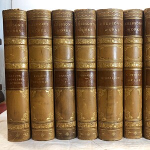 May include: A collection of antique books with brown leather covers. The spines feature gold lettering that reads "Emerson's Works" and the titles of the individual volumes. Decorative gold filigree accents the covers.