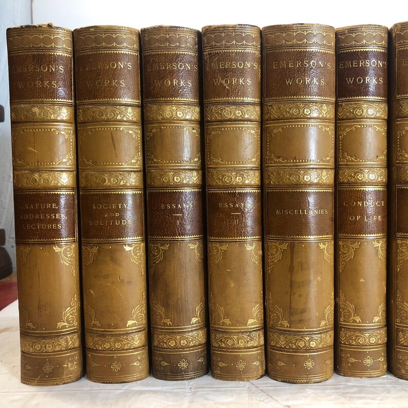 May include: A collection of antique books with brown leather covers. The spines feature gold lettering that reads "Emerson's Works" and the titles of the individual volumes. Decorative gold filigree accents the covers.