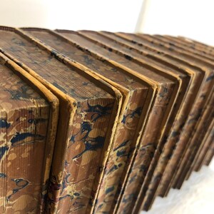 May include: A collection of antique books with marbled covers. The books are bound in brown leather with intricate patterns in shades of blue, red, and yellow. The edges of the pages are aged and yellowed, suggesting the books' age.