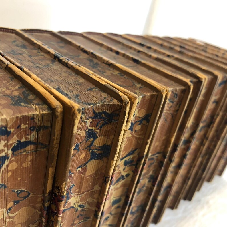 May include: A collection of antique books with marbled covers. The books are bound in brown leather with intricate patterns in shades of blue, red, and yellow. The edges of the pages are aged and yellowed, suggesting the books' age.