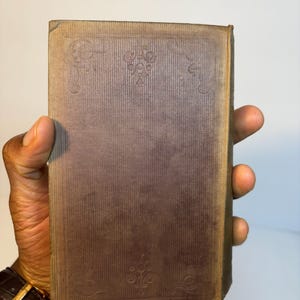 May include: A vintage book with a brown cover held in a hand. The cover has a textured, vertical striped pattern with decorative corner and center floral designs. The book's edges show wear.