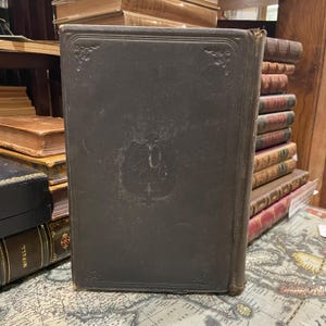 May include: A dark brown leather-bound book with a raised embossed design on the front cover. The design features a shield with the letters "A.C.G." inside.