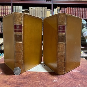 May include: Two antique leather-bound books with gold detailing and the title "Life of Peterborouh" on the spine. The book on the left is volume II and the book on the right is volume I.