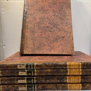 May include: A stack of antique books with brown and orange patterned covers. The spines are dark brown with gold lettering and numbers. One book stands upright on top of the stack. The books appear to be old and well-preserved.