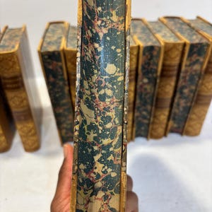 May include: A collection of antique books with aged, textured covers. The spines are a marbled pattern of teal, cream, and red. The edges are gilded, and the covers are a warm, aged tan color. The books are likely from a vintage collection.